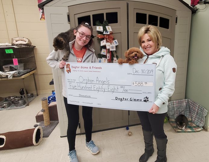 Two women holding Gizmo the dog, a cat, and a donation check for Orphan Angels 