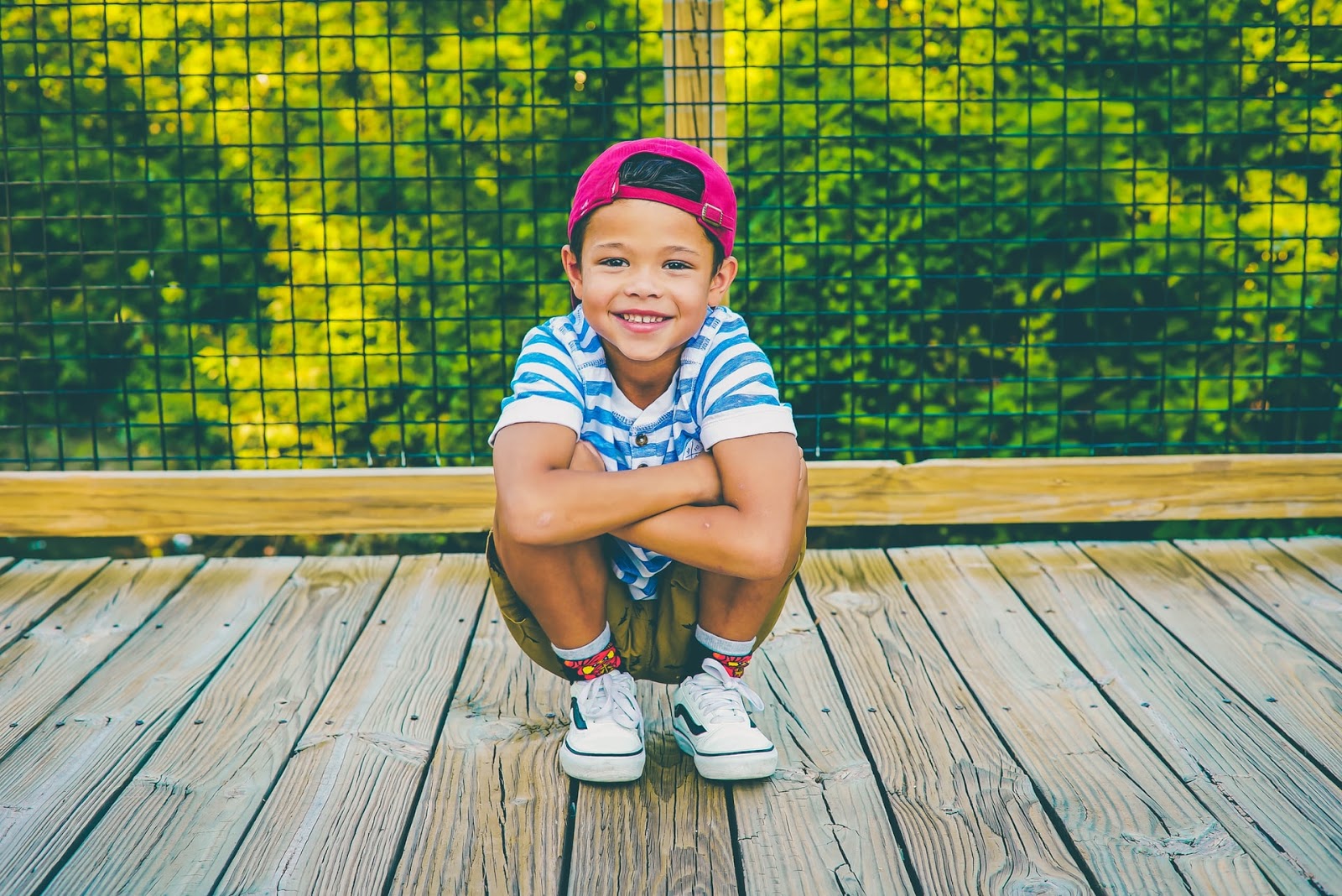 A young boy in a baseball cap, crouching on a wooden bridge 